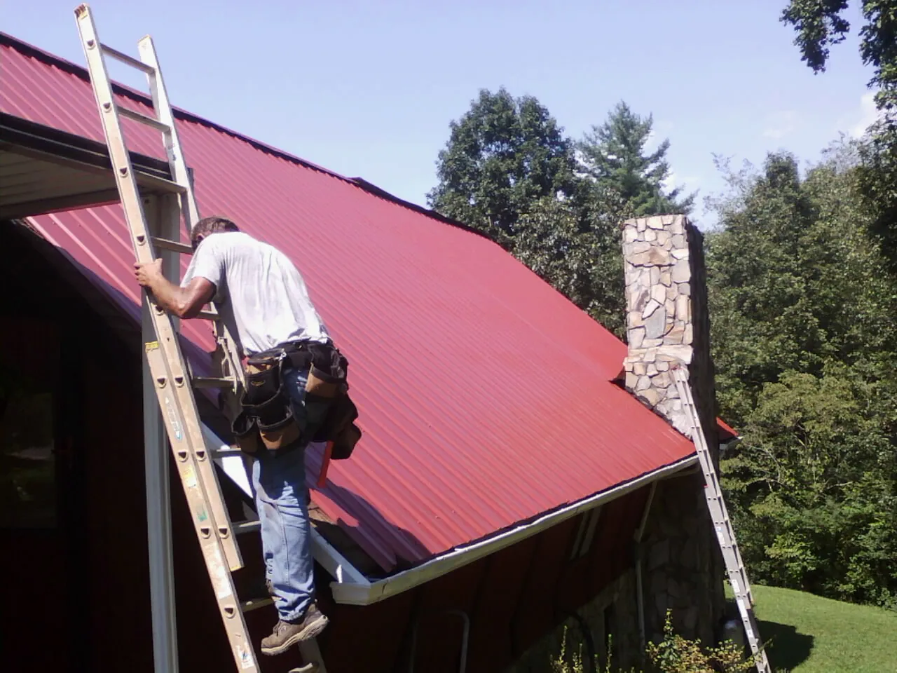 Roofer on a ladder beside a red metal roof and stone chimney — Soffit & Fascia Repair by Roofing Brothers in Raleigh